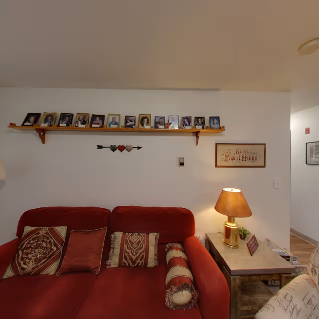 A cozy living room area with a red couch adorned with patterned pillows and a bolster pillow. Above the couch is a wooden shelf displaying framed photographs. To the right of the couch is a side table with a lamp, a small plant, and a framed sign that reads 'Home Sweet Home'. The walls are white and the floor is wooden.