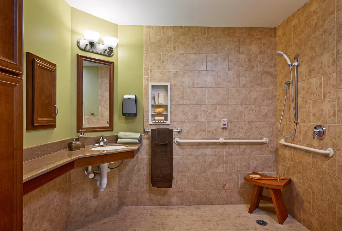 Accessible bathroom with beige tiled walls and floor, featuring a walk-in shower with a handheld showerhead, grab bars, a wooden shower stool, a sink with a granite countertop, a mirror, a wall-mounted soap dispenser, and towels.