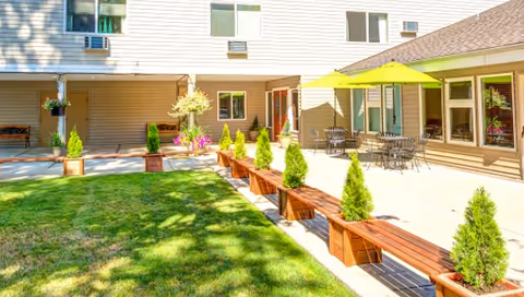 Sunny courtyard with wooden benches and small potted evergreens, patio tables with green umbrellas, and a multi-story building facade.