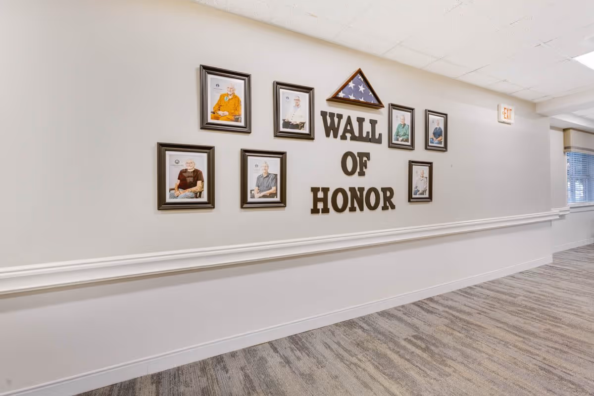 A hallway wall display titled 'Wall of Honor' featuring framed photographs of elderly individuals and a folded American flag in a triangular case above the text. The hallway has light-colored walls, a carpeted floor, and an exit sign near the ceiling.