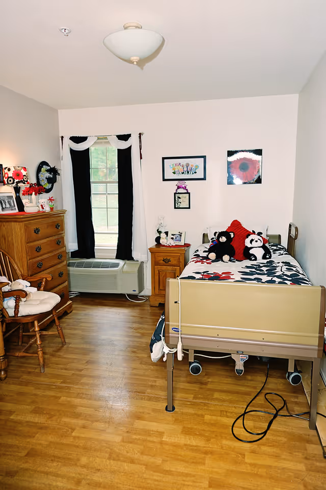 A senior care facility bedroom with a hospital-style bed featuring a floral bedspread and stuffed animals. The room has wooden flooring, a wooden dresser with framed photos and decorations, a wooden rocking chair with a cushion, a small wooden nightstand with a lamp and picture frame, and a window with black and white curtains. The walls are decorated with framed artwork.