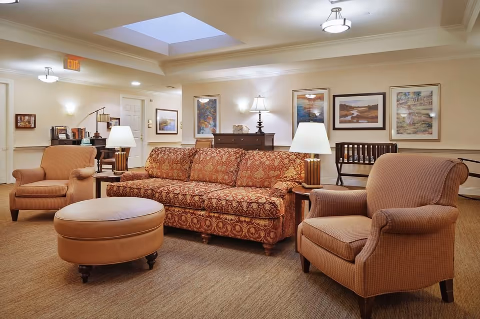 A cozy living room area with a patterned red sofa, two beige armchairs, and a round beige ottoman. The room is well-lit with table lamps on side tables, wall sconces, and ceiling lights. There are framed paintings on the walls and a skylight in the ceiling. A wooden crib and a dresser are visible in the background.