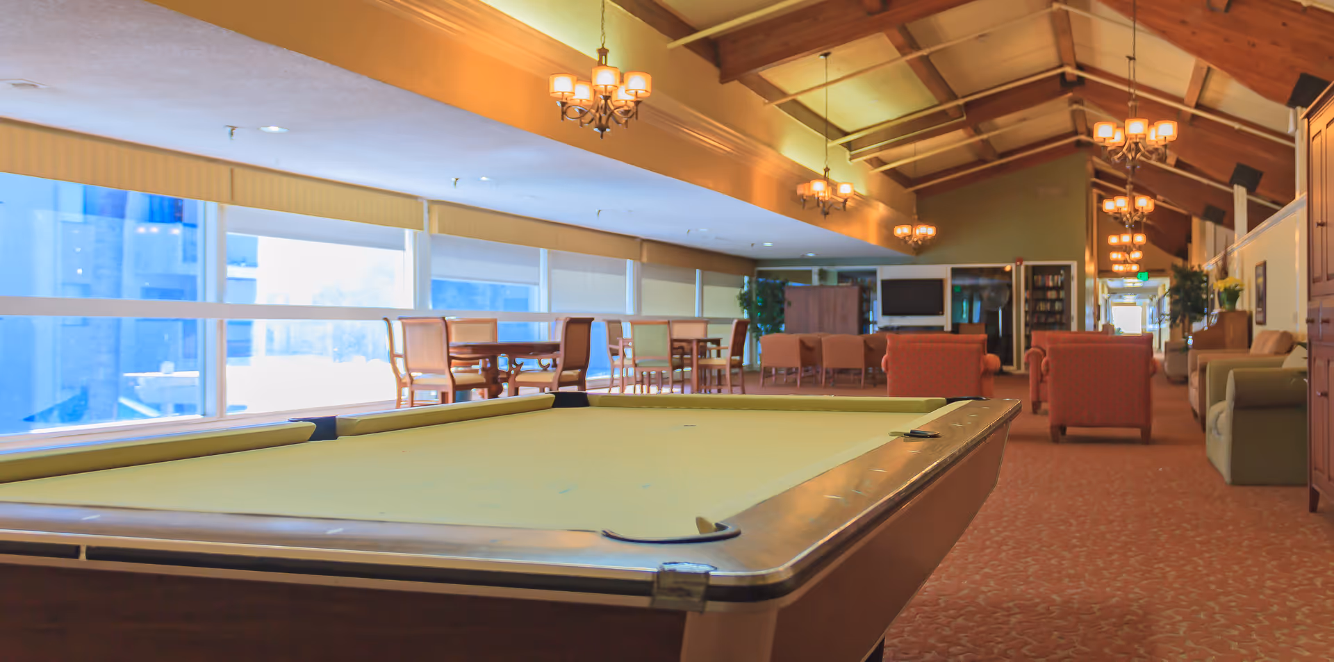 A spacious recreational room with a pool table in the foreground, several seating areas with armchairs and sofas, wooden tables with chairs near large windows, and chandeliers hanging from a high wooden ceiling.