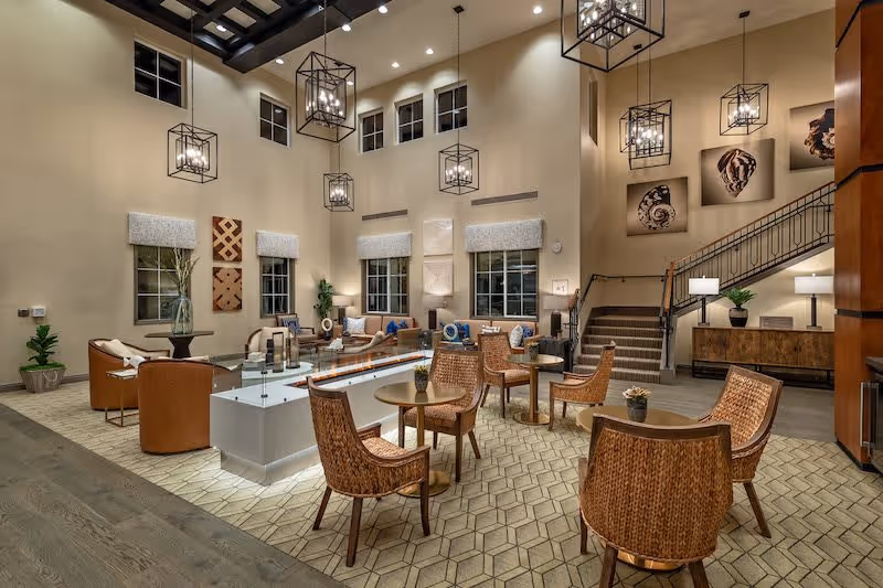 A spacious and well-lit senior living facility common area with high ceilings and large windows. The room features multiple seating arrangements including wicker chairs around small round tables and cushioned armchairs around a modern rectangular fireplace. Decorative hanging light fixtures and wall art with seashell designs adorn the space. A staircase with a metal railing leads to an upper level, and there are plants and lamps adding to the cozy ambiance.