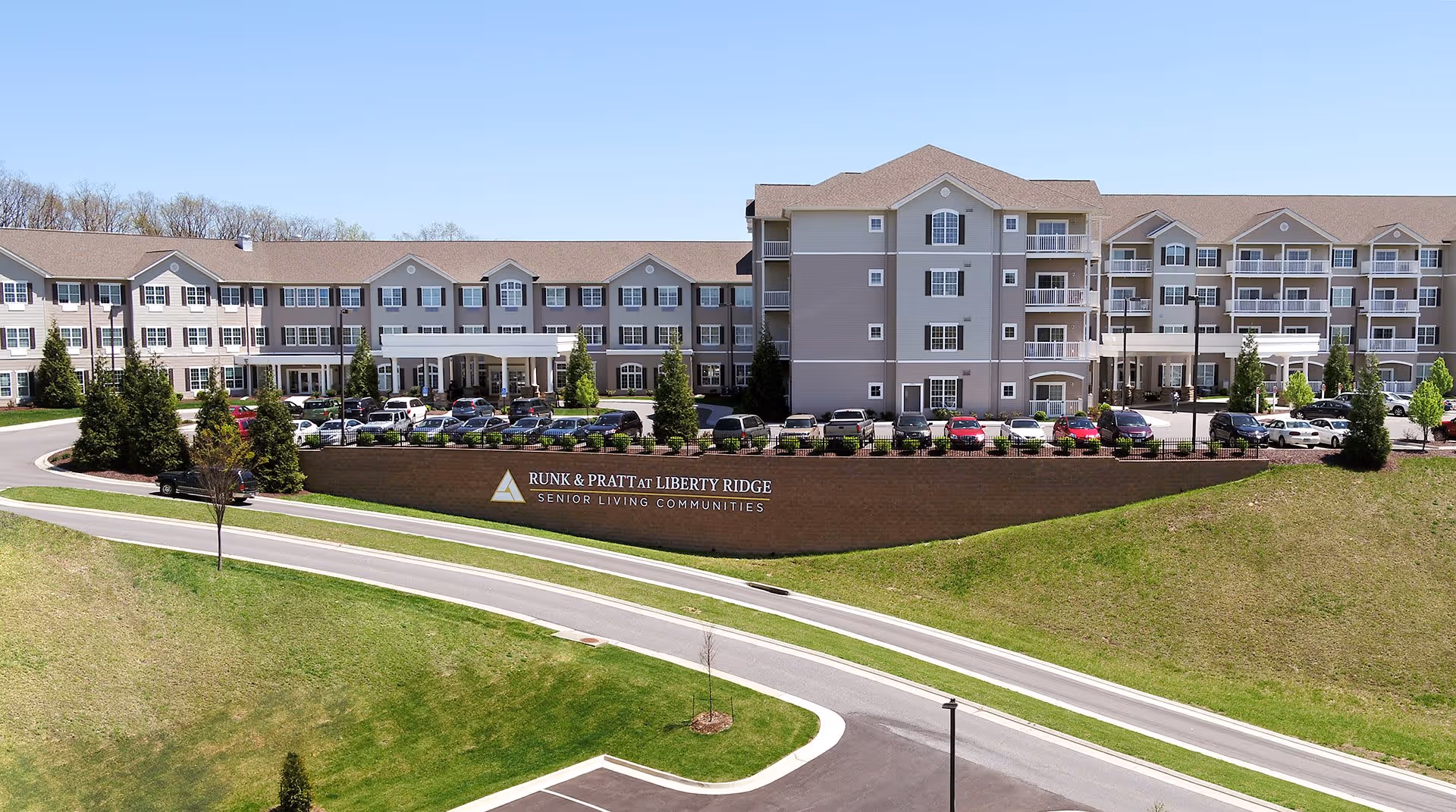 Exterior view of Runk & Pratt at Liberty Ridge senior living community showing a large multi-story building with balconies, numerous parked cars, a curved driveway, and a grassy area in the foreground under a clear blue sky.