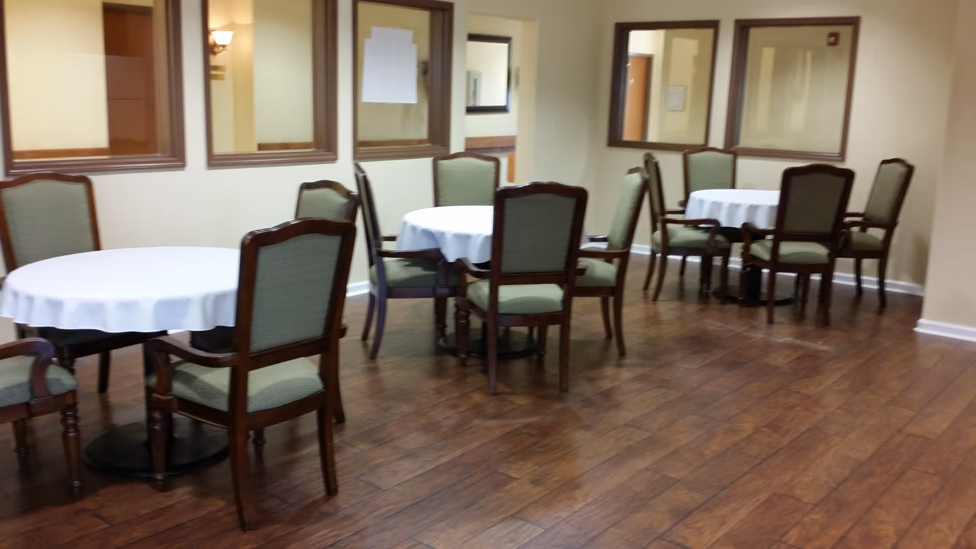 Interior view of a dining area with round tables covered with white tablecloths and surrounded by wooden chairs with green upholstery. The room has wooden flooring and beige walls with several windows looking into adjacent rooms.