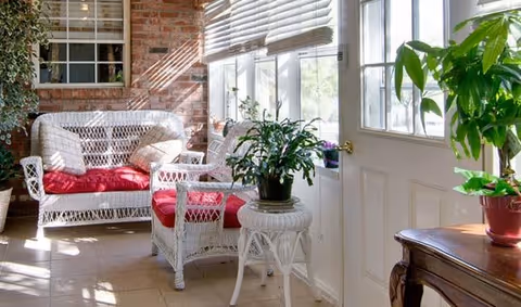 A sunlit indoor seating area with white wicker furniture including a loveseat and chair with red cushions, a small white wicker table holding a potted plant, and a wooden table with another potted plant near a white door and windows with blinds.