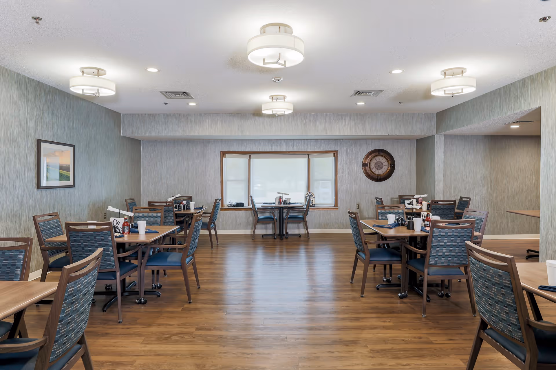 A dining room in an assisted living facility with several wooden tables and chairs arranged neatly. The room has wooden flooring, light gray walls, and a large window at the far end. Ceiling lights provide bright illumination, and there is a large wall clock and framed artwork on the walls.