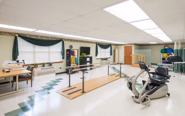 A bright physical therapy room with parallel walking bars, a recumbent exercise bike, weight rack, and other rehabilitation equipment.