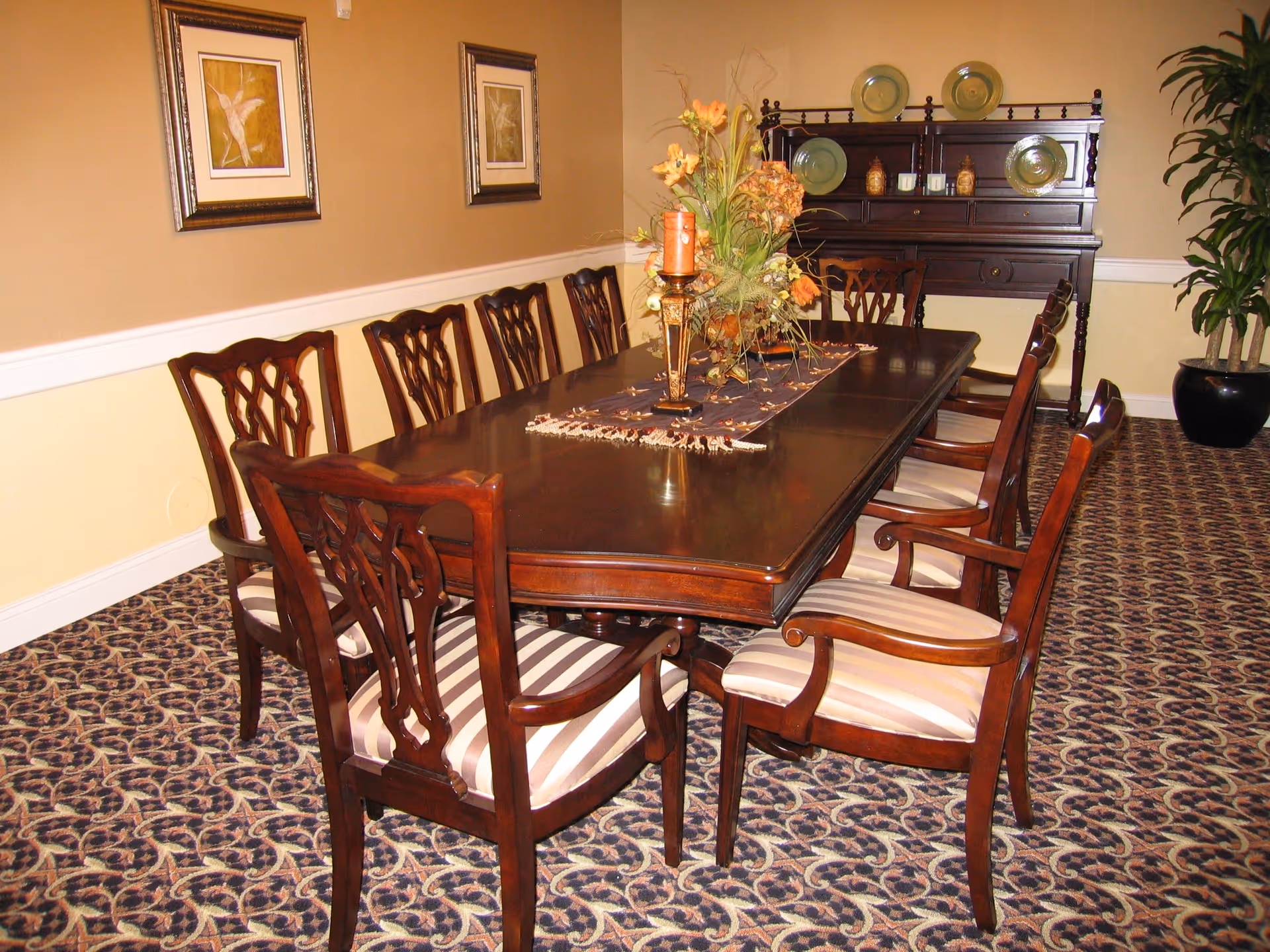 A formal dining room with a long dark wooden table surrounded by eight matching chairs with striped cushions. The table is decorated with a floral centerpiece and a candle. The room has patterned carpet, beige walls with white wainscoting, two framed pictures on the wall, a dark wooden sideboard with decorative plates, and a large potted plant in the corner.