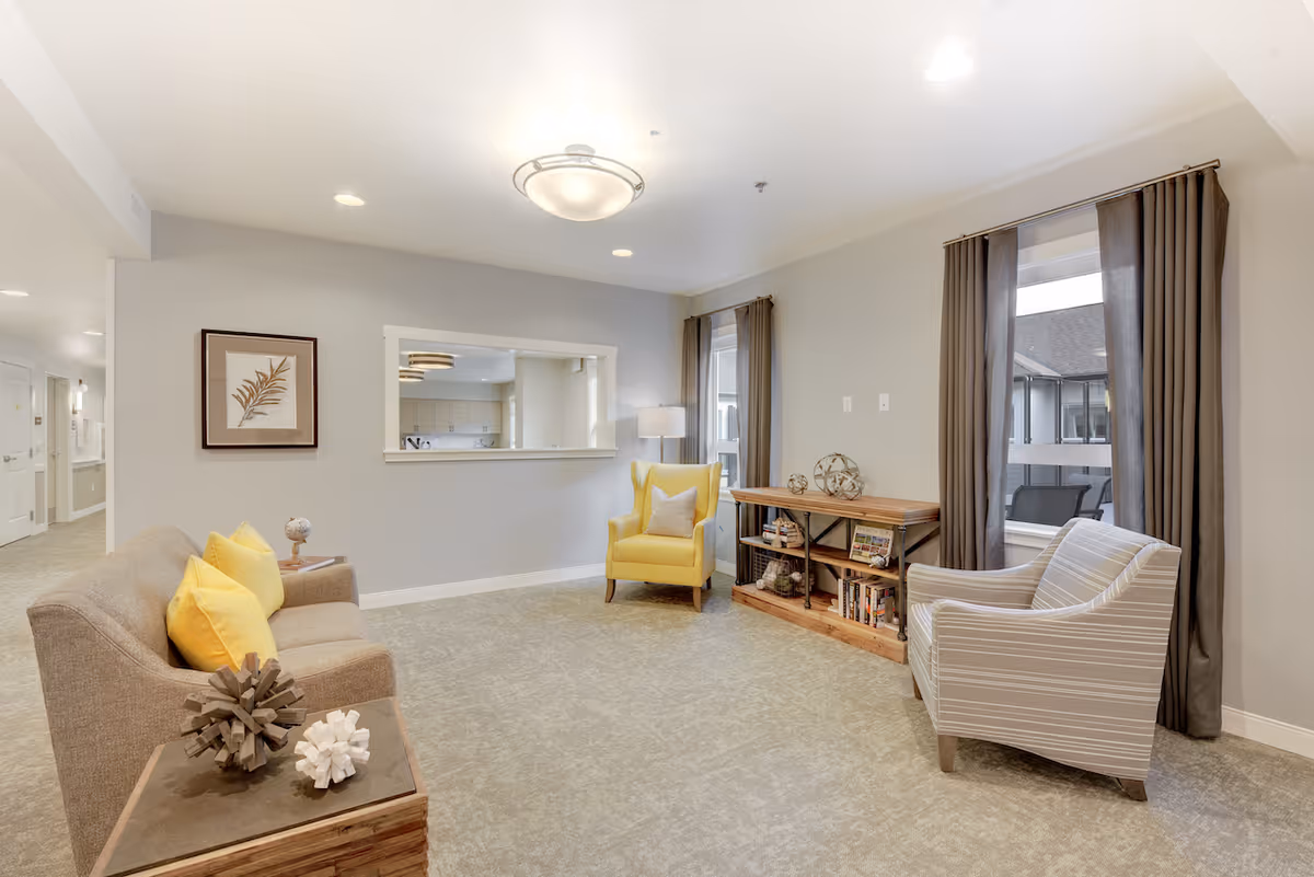 Well-lit communal living room with sofas, armchairs (including a yellow chair), a bookcase, and decorative accents.