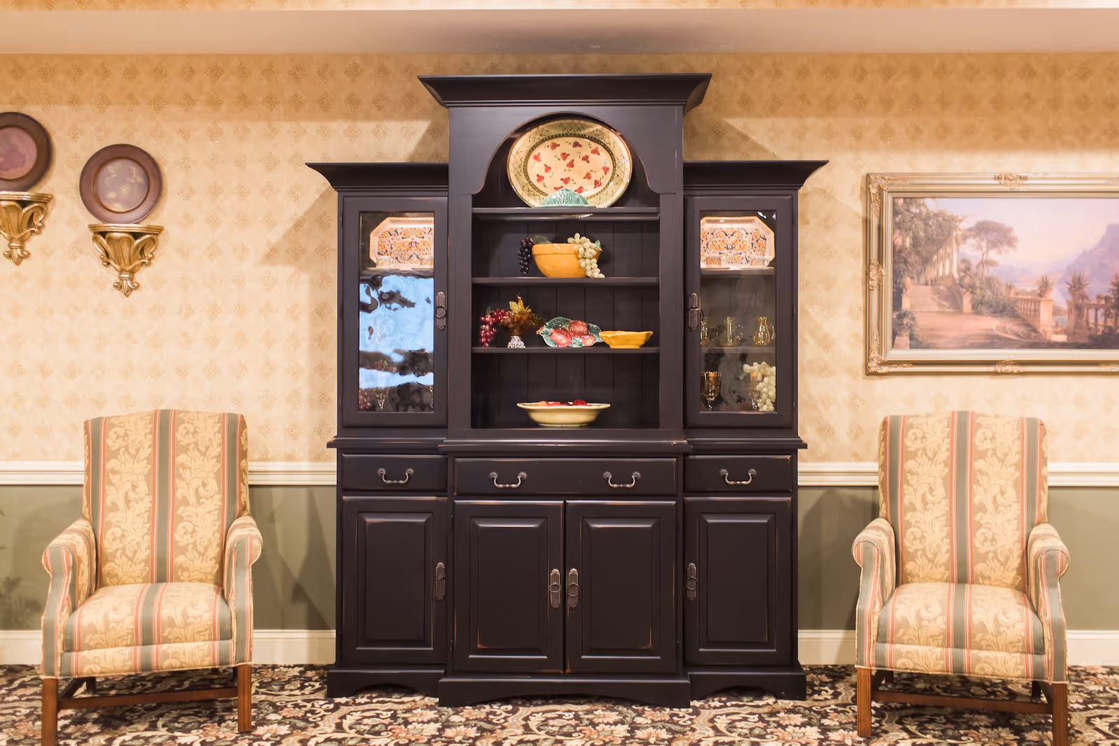 A traditional living room area featuring a dark wooden cabinet with decorative plates and artificial fruit on display. Two upholstered armchairs with a floral and striped pattern are positioned on either side of the cabinet. The walls have a patterned wallpaper with a chair rail and a framed landscape painting on the right side.
