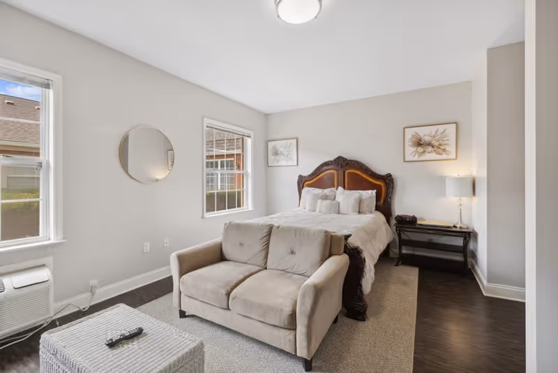 A cozy bedroom featuring a large bed with an ornate wooden headboard, multiple pillows, and two framed floral artworks on the wall above. In front of the bed is a beige loveseat sofa and a woven ottoman with a remote control on top. The room has two windows letting in natural light, a nightstand with a lamp and phone, and dark wood flooring with a light area rug.