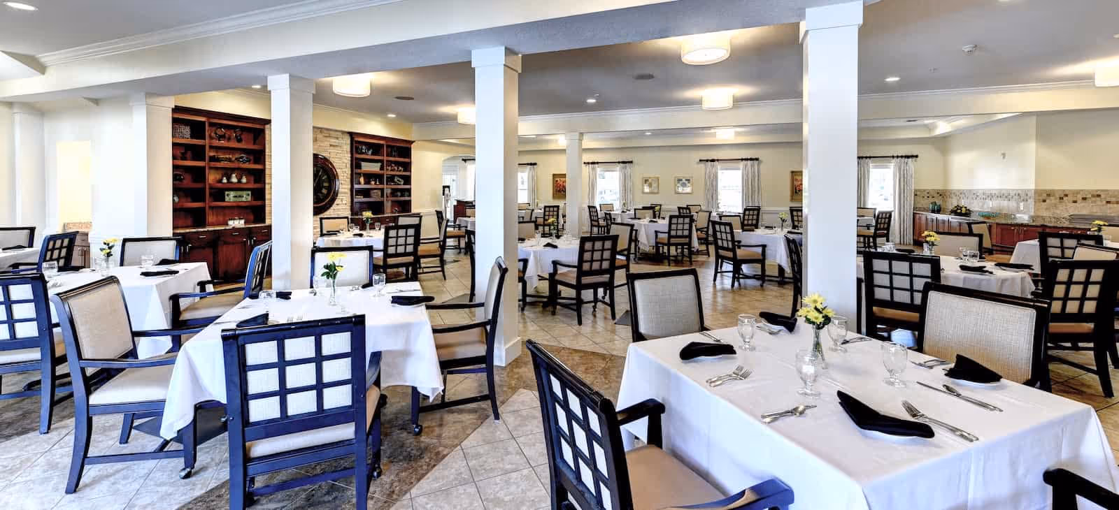 A spacious dining room with multiple tables covered in white tablecloths, each set with black napkins, silverware, and glassware. The room features tiled floors, wooden chairs with cushioned seats, white pillars, and built-in wooden shelves along one wall. Soft lighting and windows with curtains provide a bright and inviting atmosphere.