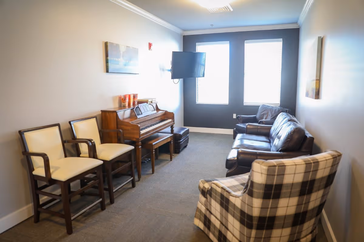 A cozy living room area with a piano against the left wall, two cream-colored chairs next to it, a plaid armchair in the foreground, and two dark leather sofas near two large windows with blinds. A flat-screen TV is mounted on the wall between the windows, and there are paintings on the walls.