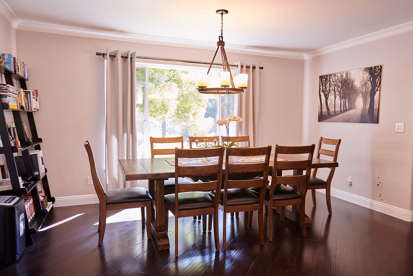 Sunlit dining room with a wooden table and six chairs beneath a hanging chandelier, a large window with curtains, a bookshelf to the left, and wall art on the right.