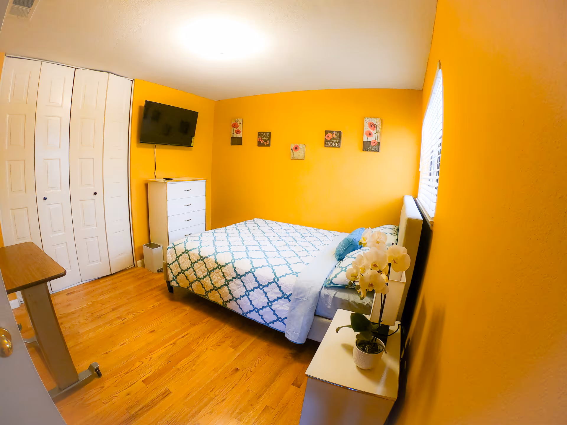 A cozy bedroom with bright yellow walls, a bed with a white and blue patterned bedspread, a white dresser with a flat-screen TV mounted above it, a small white nightstand with a potted orchid, and decorative wall art featuring flowers and inspirational words.