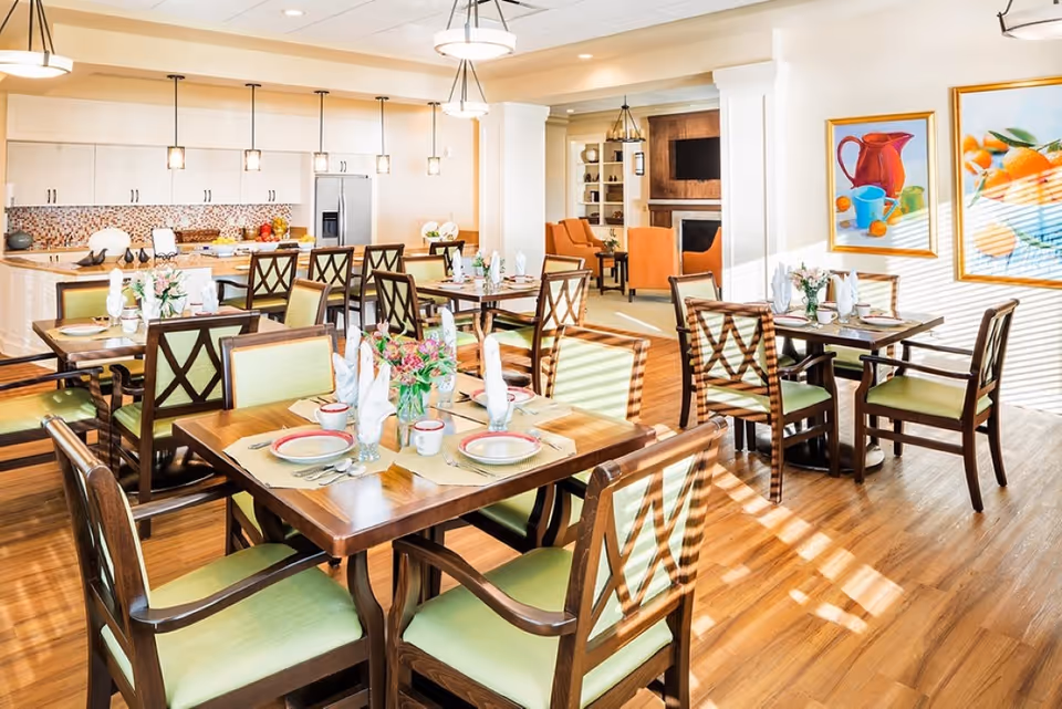 Bright and spacious dining area with multiple wooden tables and green cushioned chairs set with plates, cups, and napkins. The room features a kitchen area with white cabinets, a refrigerator, and pendant lights hanging from the ceiling. In the background, there is a cozy sitting area with orange chairs and a fireplace, along with colorful framed artwork on the walls.