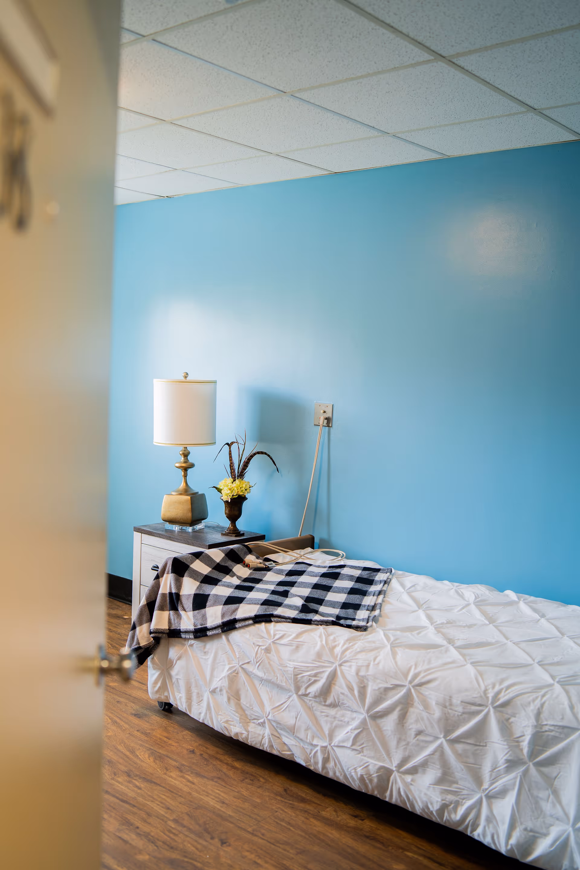 Small bedroom with a single bed, nightstand holding a lamp and flowers against a blue wall.