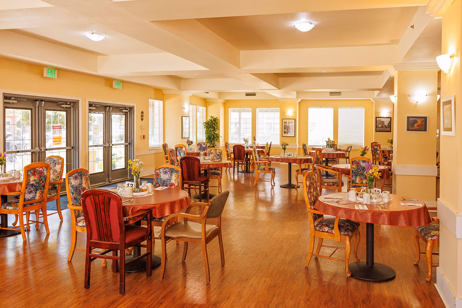 A bright and spacious dining room with multiple round tables covered with red tablecloths. Each table is set with cups, utensils, and small flower arrangements. The chairs around the tables have floral upholstery and wooden frames. Large windows and glass doors allow natural light to fill the room, and the walls are painted a soft yellow. The floor is wooden, and framed pictures hang on the walls.