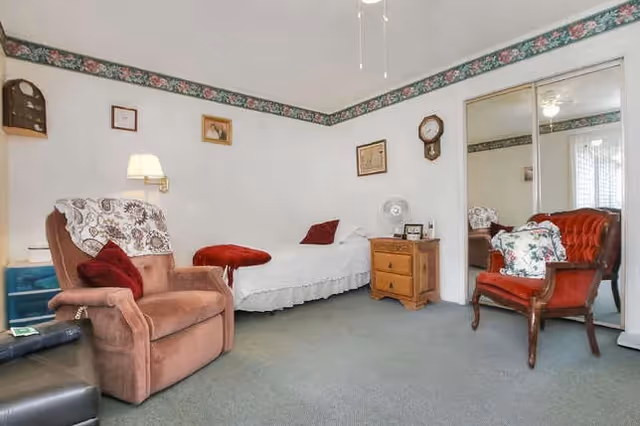 A cozy room with a single bed covered in a white bedspread, a wooden nightstand with a clock and fan, a brown recliner chair with a white lace cover and red pillow, a red cushioned armchair with a floral pillow, and mirrored closet doors. The walls are white with a floral border near the ceiling, and there are several framed pictures and a wall clock hanging on the walls.