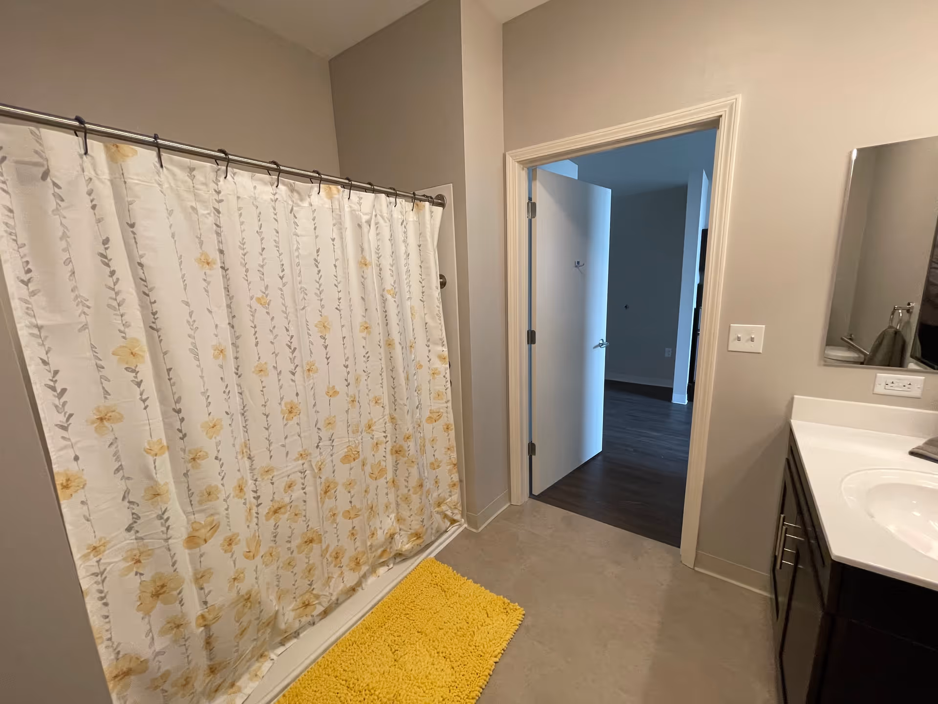 Bathroom with a shower area covered by a floral-patterned shower curtain, a yellow bath mat on the floor, a white door open to an adjacent room with dark wood flooring, and a vanity with a sink and mirror on the right side.