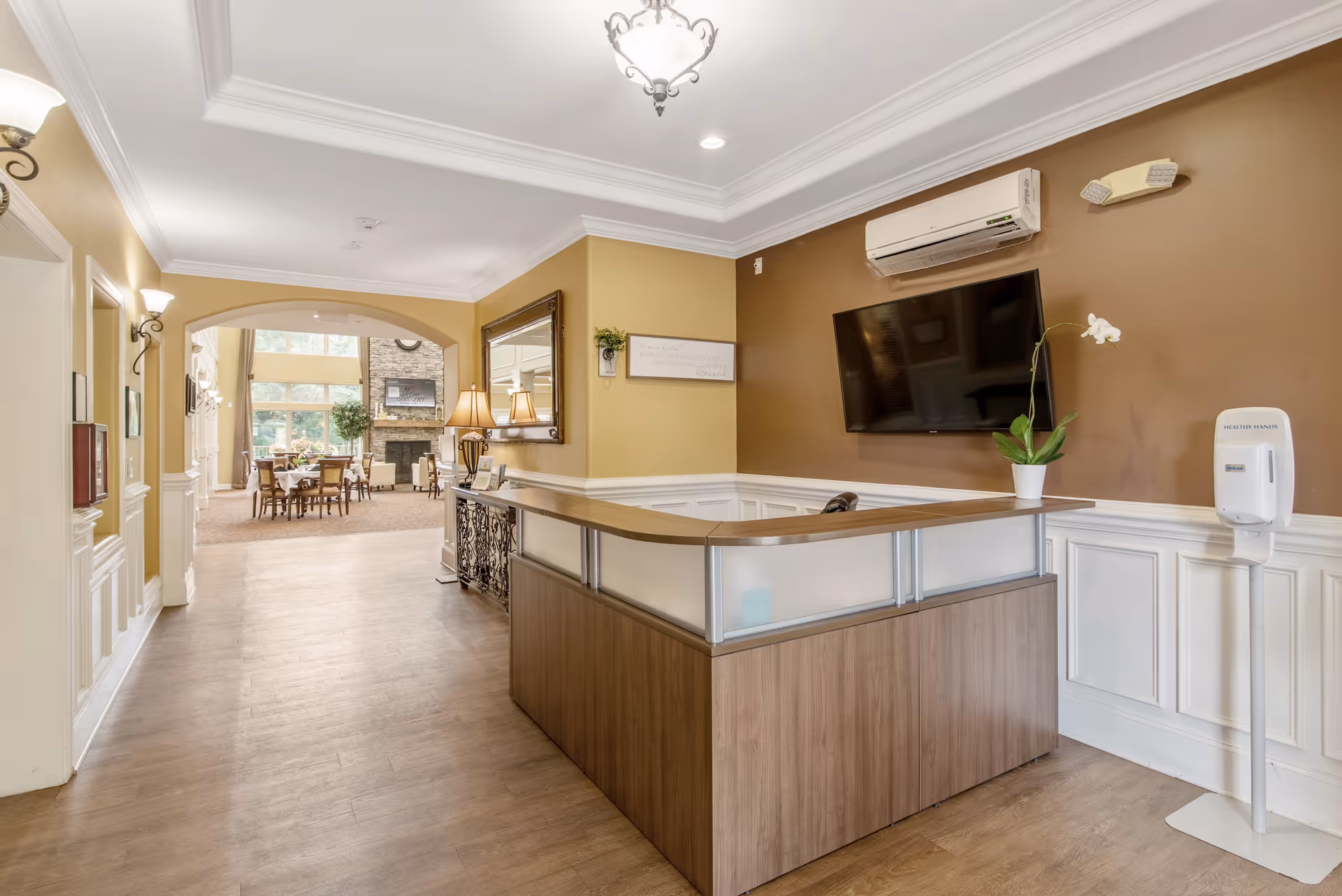 Reception area with a wooden desk and a potted orchid plant, a wall-mounted TV, and a hand sanitizer dispenser. The hallway leads to a bright common area with tables, chairs, large windows, and a stone fireplace.