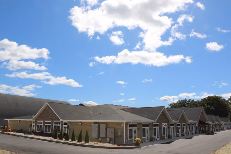 Exterior view of a single-story senior living facility building with multiple entrances, a sloped roof, and a clear blue sky with scattered clouds above.