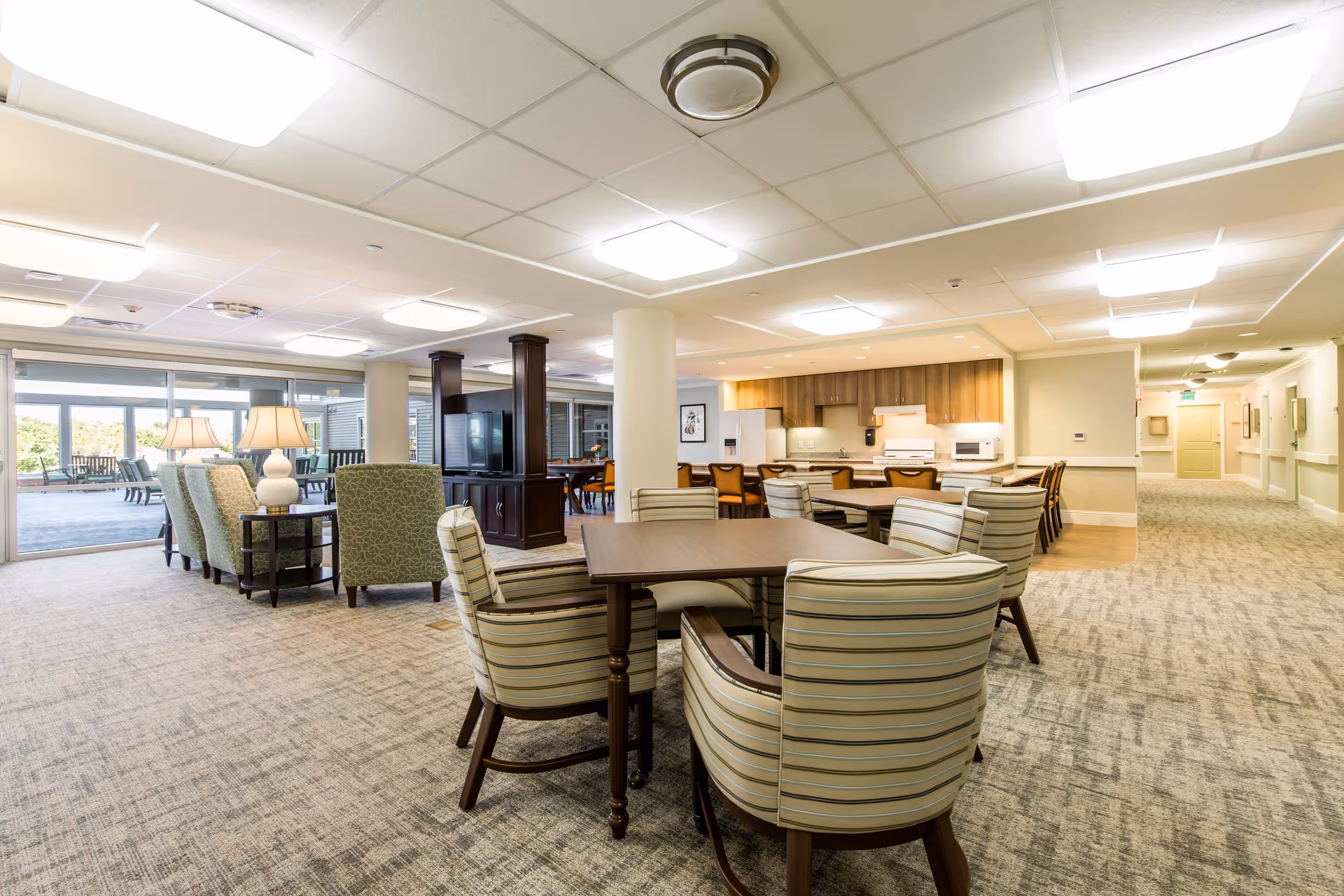 A spacious and well-lit common area in a senior living facility featuring multiple seating arrangements with striped and patterned upholstered chairs around wooden tables. The room includes a kitchen area with wooden cabinets, white appliances, and a countertop with chairs. Large windows and glass doors in the background lead to an outdoor patio with additional seating. The carpeted floor and ceiling lights create a warm and inviting atmosphere.