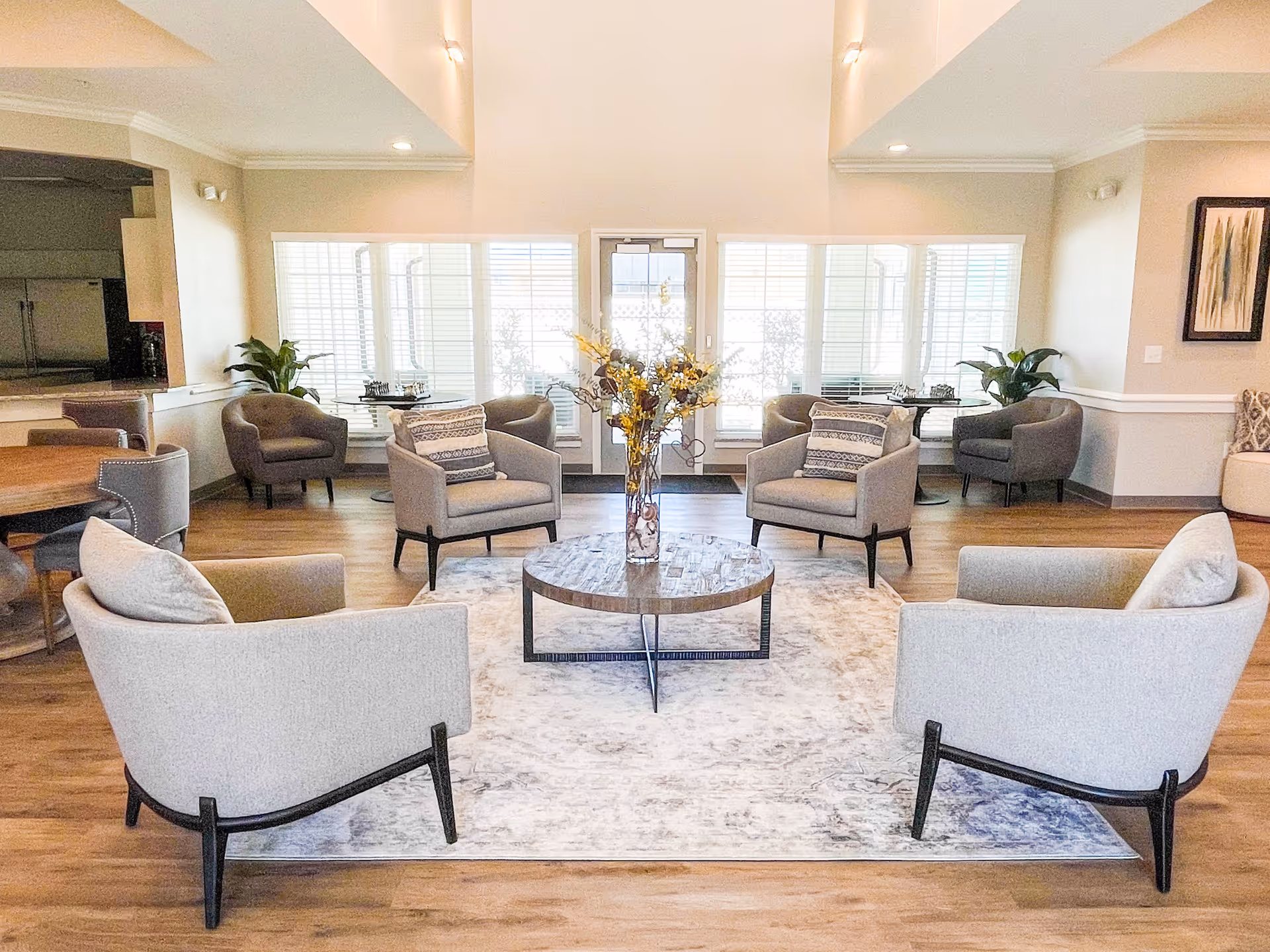 Bright, modern senior living common room with multiple armchairs arranged around a central coffee table and large windows in the background.