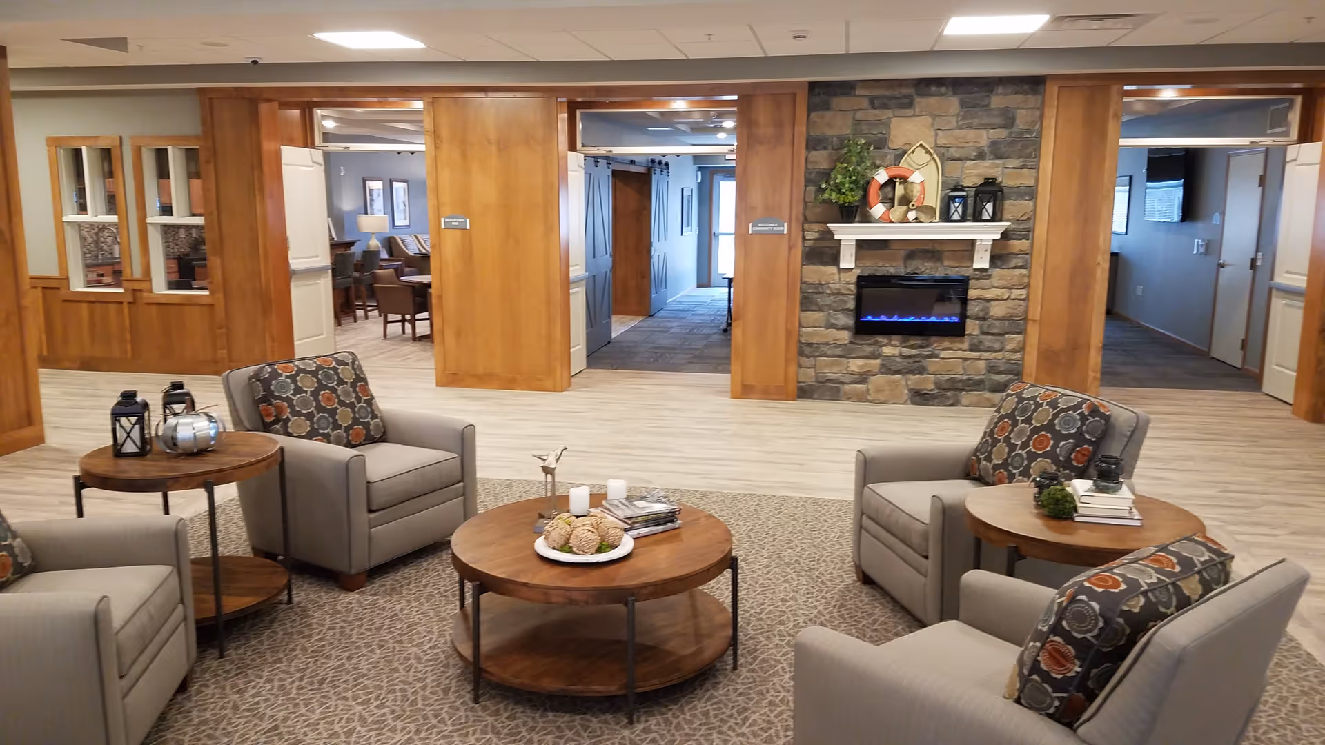 A cozy senior living common area with four gray armchairs arranged around a round wooden coffee table. The chairs have patterned cushions. There are two side tables with decorative items including lanterns and books. The room features a stone wall with a modern electric fireplace and nautical-themed decorations on the mantel. The background shows wooden paneling, open doorways leading to other rooms, and a carpeted floor area transitioning to wood flooring.