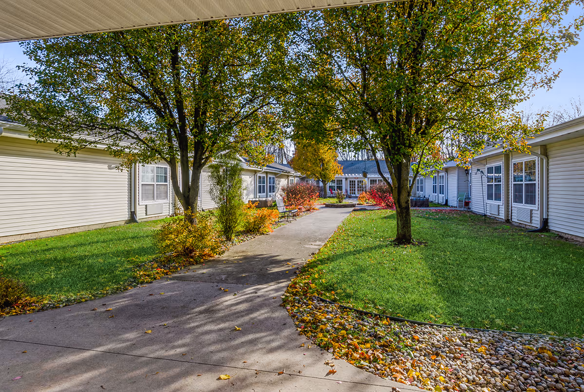 A paved walkway flanked by green lawns, trees with autumn leaves, and shrubs leads to a building with white siding and multiple windows under a clear blue sky.