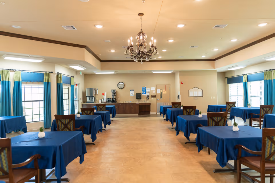 A dining room with multiple tables covered in blue tablecloths, each with a small decorative plant. The room has large windows with blue and green curtains, a chandelier hanging from the ceiling, and a counter area with coffee machines and a clock on the wall.