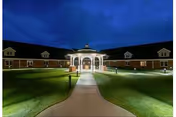 Nighttime exterior view of a single-story building with a well-lit white gazebo at the center, surrounded by a curved walkway and neatly maintained grass lawn under a dark blue sky.