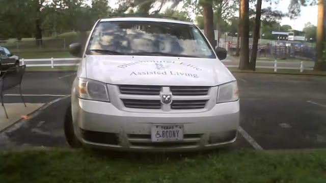 Front view of a white Dodge vehicle parked in a parking lot with trees and a playground in the background. The vehicle has 'Bluebonnet Elite Assisted Living' written on the hood and a Texas license plate with a handicap symbol.