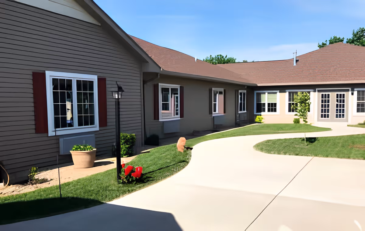 Exterior view of a senior living facility with a curved concrete pathway, green lawn, small plants, and a building with multiple windows and a brown roof under a clear blue sky.