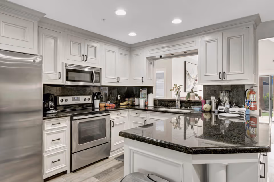 Modern kitchen with white cabinetry, black granite countertops, stainless steel refrigerator, stove, and microwave. The kitchen features a coffee maker, blender, and various kitchen utensils on the counters. There is a pass-through window opening to another room with a piece of wall art visible.