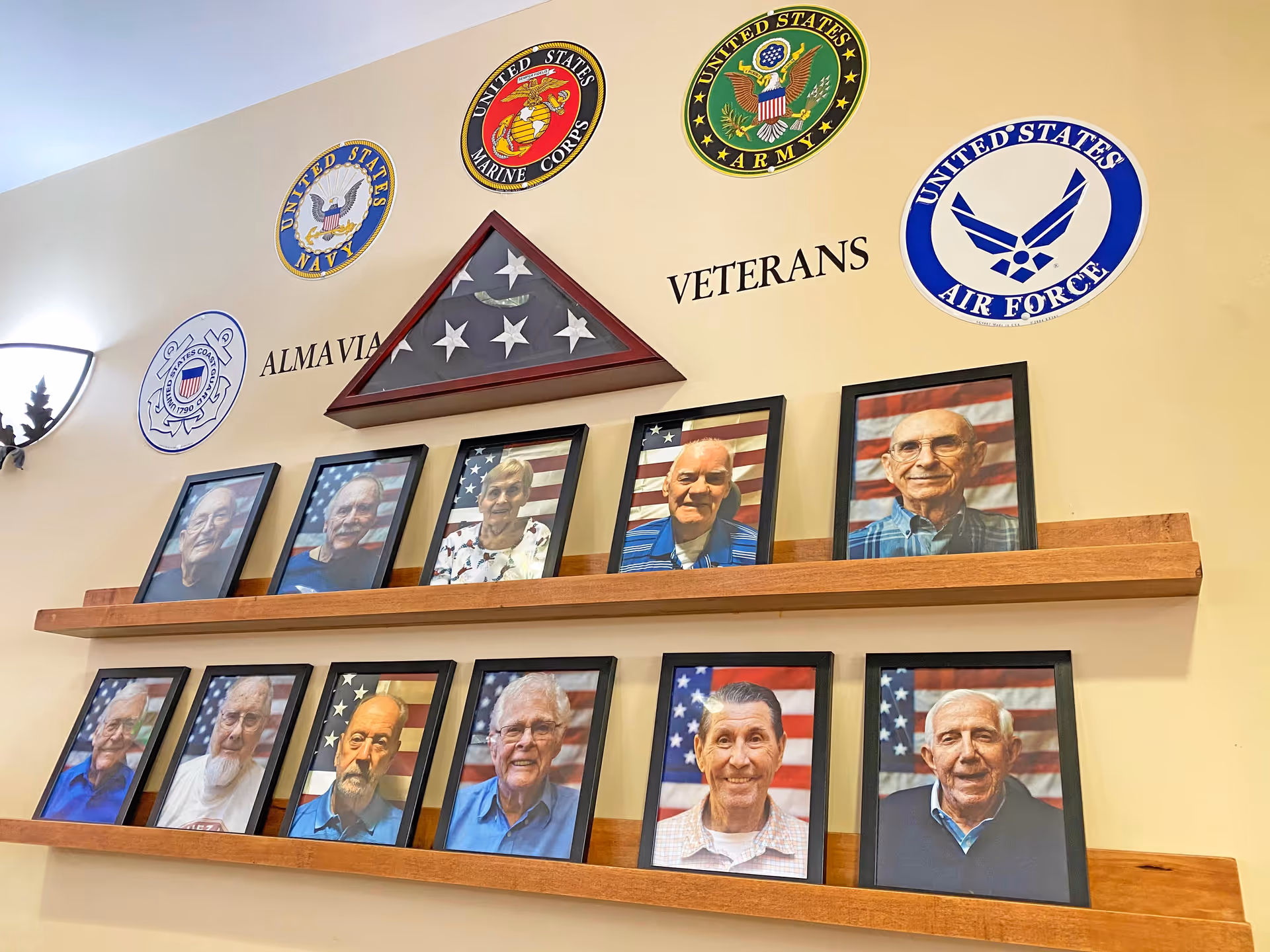 A wall display at AlmaVia of Camarillo honoring veterans. The display features framed portraits of elderly veterans arranged on two wooden shelves. Above the portraits are emblems of the United States Navy, Marine Corps, Army, Coast Guard, and Air Force, along with a folded American flag in a triangular case. The words 'ALMAVIA VETERANS' are written on the wall between the emblems and the portraits.