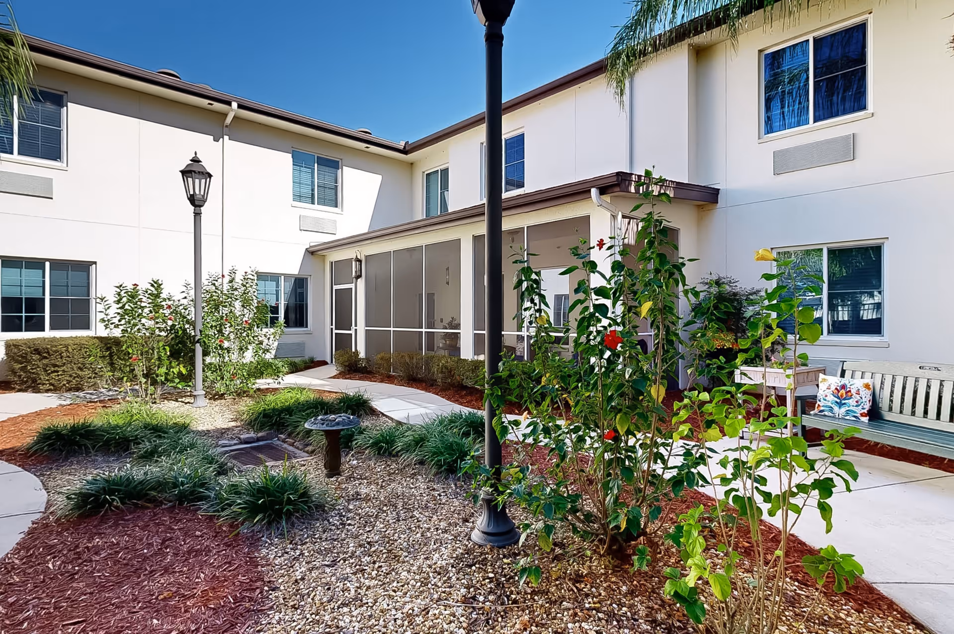 Outdoor courtyard area of a senior living facility with a garden featuring various plants and flowers, a birdbath, a paved walkway, a bench with a decorative pillow, and a two-story building with multiple windows in the background under a clear blue sky.
