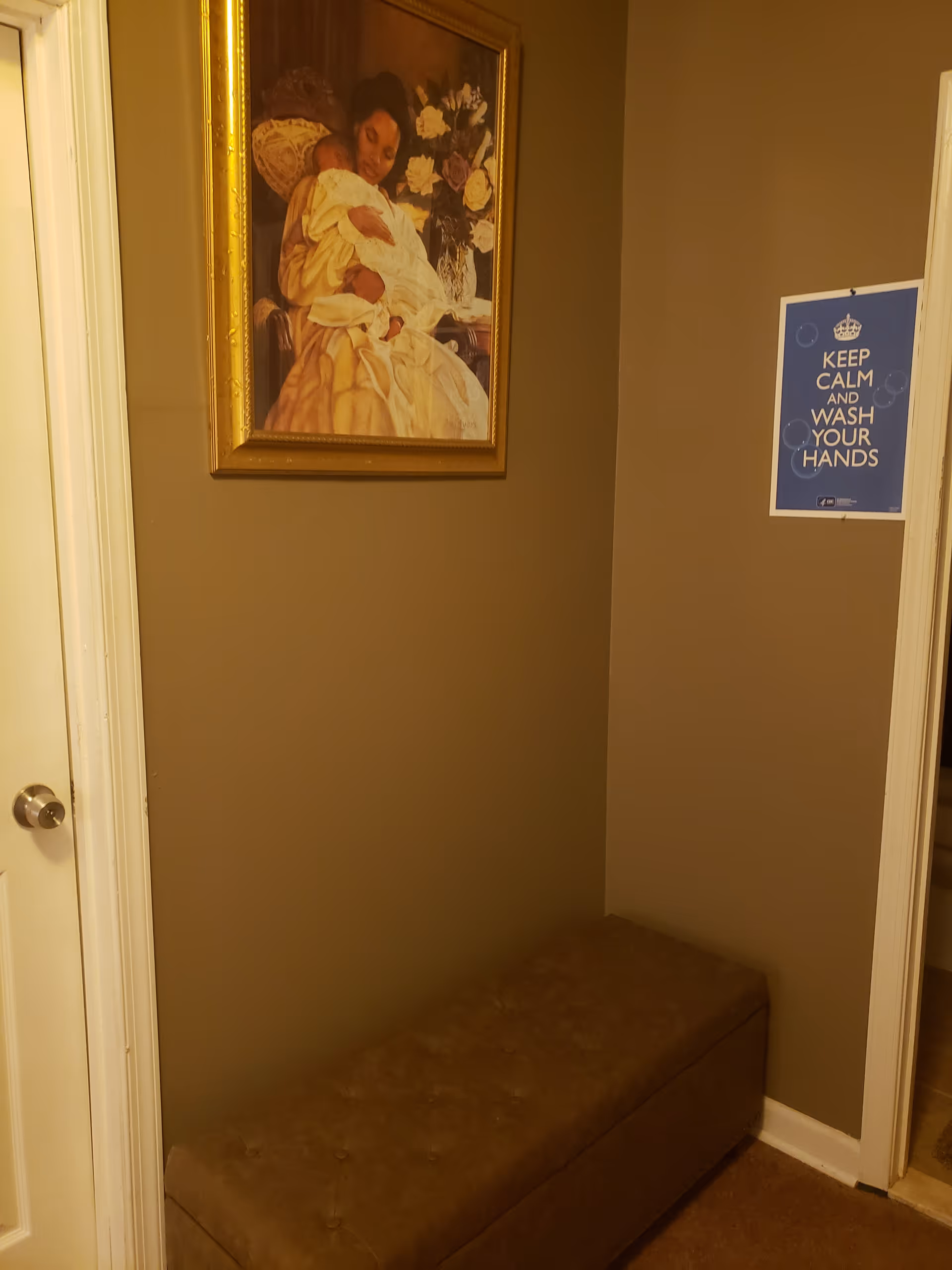A small corner of a room with brown walls featuring a cushioned bench, a framed painting of a woman holding a baby, and a blue sign on the wall that reads 'KEEP CALM AND WASH YOUR HANDS'. A white door with a silver doorknob is partially visible on the left.