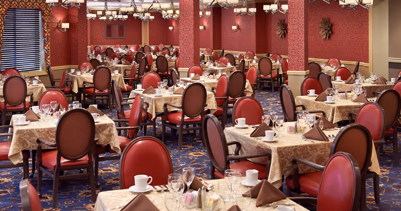 A spacious dining room with multiple tables covered in beige tablecloths, each set with white cups, glasses, silverware, and brown folded napkins. The chairs have red cushioned seats and backs with dark wooden frames. The walls are decorated with red patterned wallpaper and light fixtures, and the carpet has a blue and gold floral pattern.