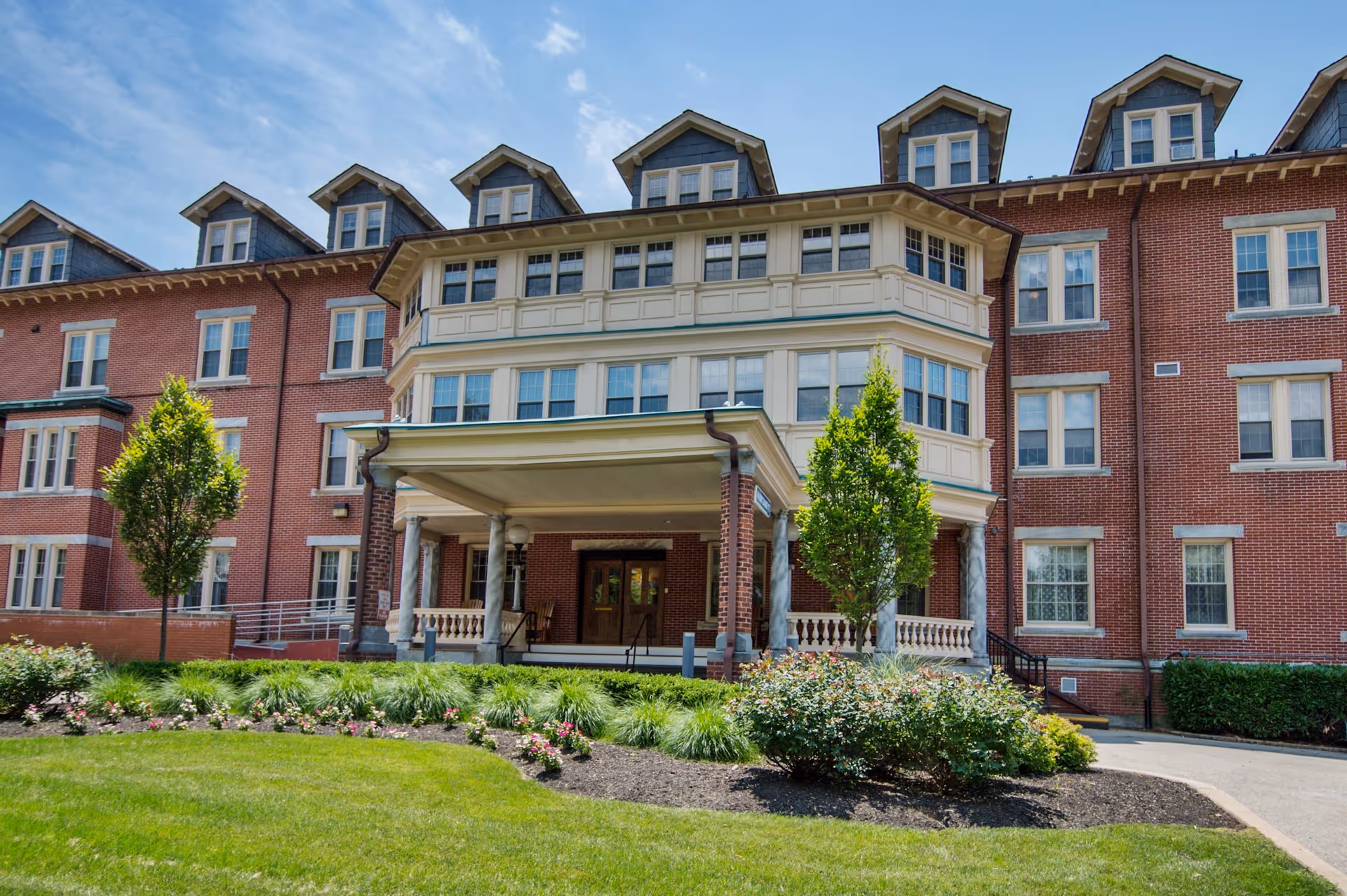 Front exterior view of a large, multi-story brick building with multiple windows and dormer windows on the roof. The building has a covered entrance supported by columns, surrounded by well-maintained landscaping including green grass, bushes, and small trees under a partly cloudy blue sky.
