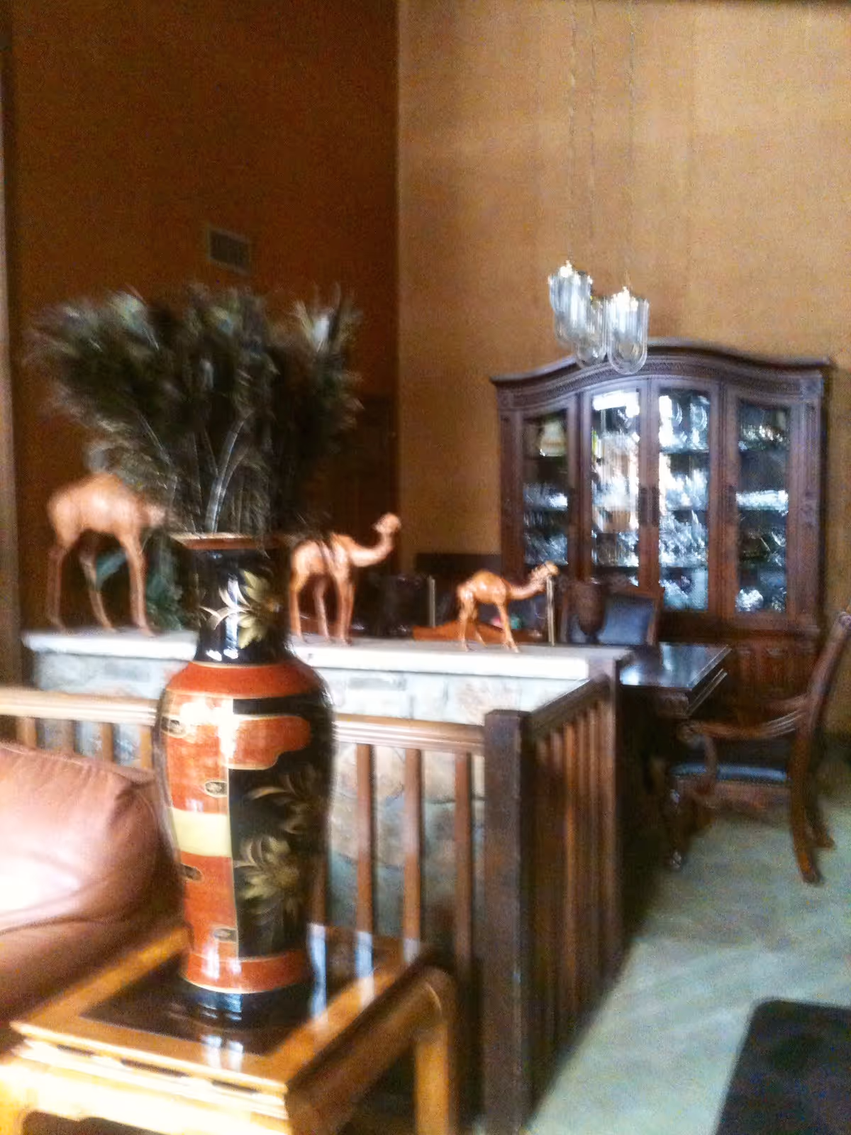 Interior view of a living room area featuring a large decorative vase with tall feathers on a wooden side table, three wooden camel figurines on a stone ledge, a wooden dining table with chairs, and a glass-fronted wooden cabinet filled with glassware. The walls are painted in warm tones and a chandelier hangs from the ceiling.