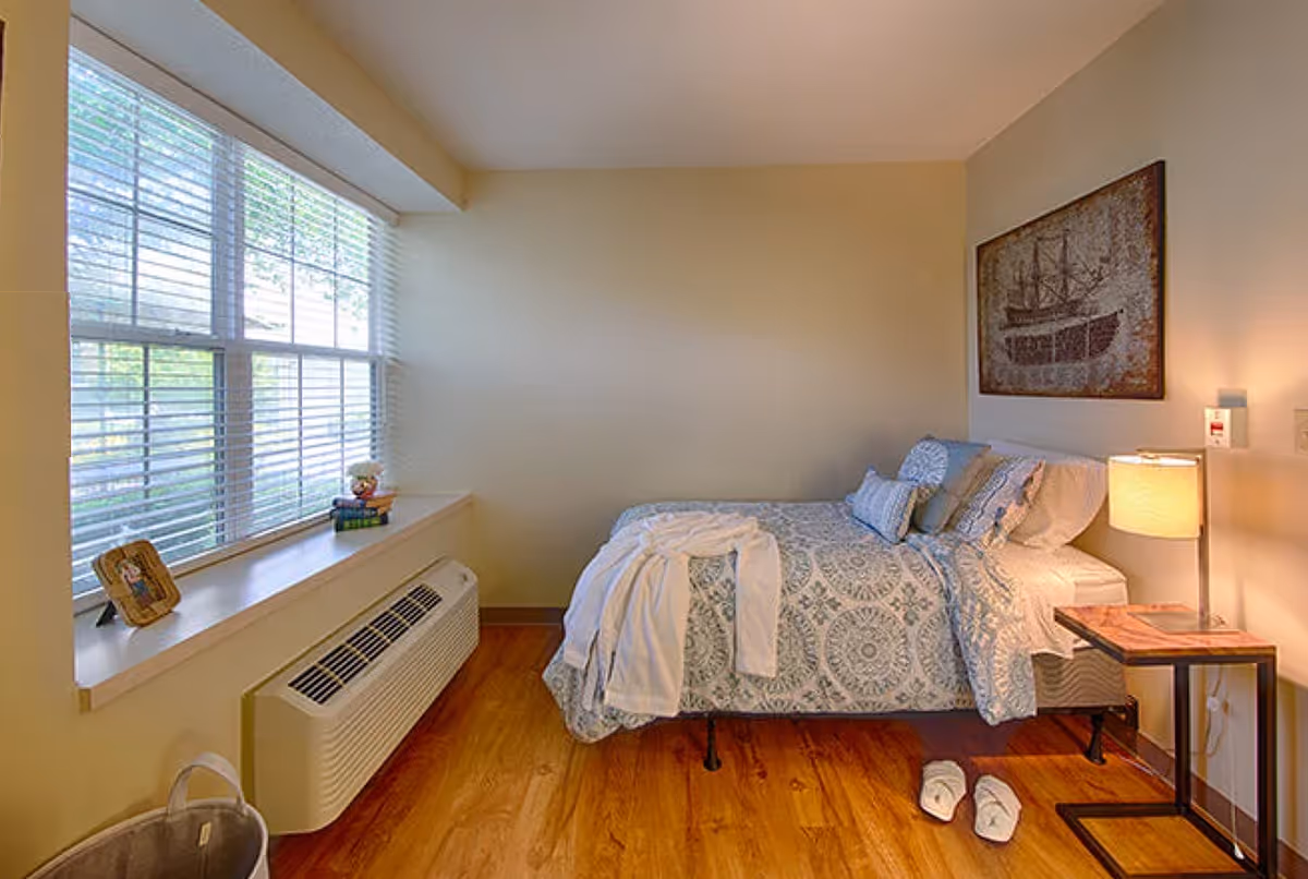 A cozy bedroom with a single bed covered in a patterned blue and white bedspread, a white robe laid out on the bed, and slippers on the wooden floor next to it. A small side table with a lamp is beside the bed, and a framed picture of a ship hangs on the wall above. A large window with blinds lets in natural light, and a heating/cooling unit is below the window.