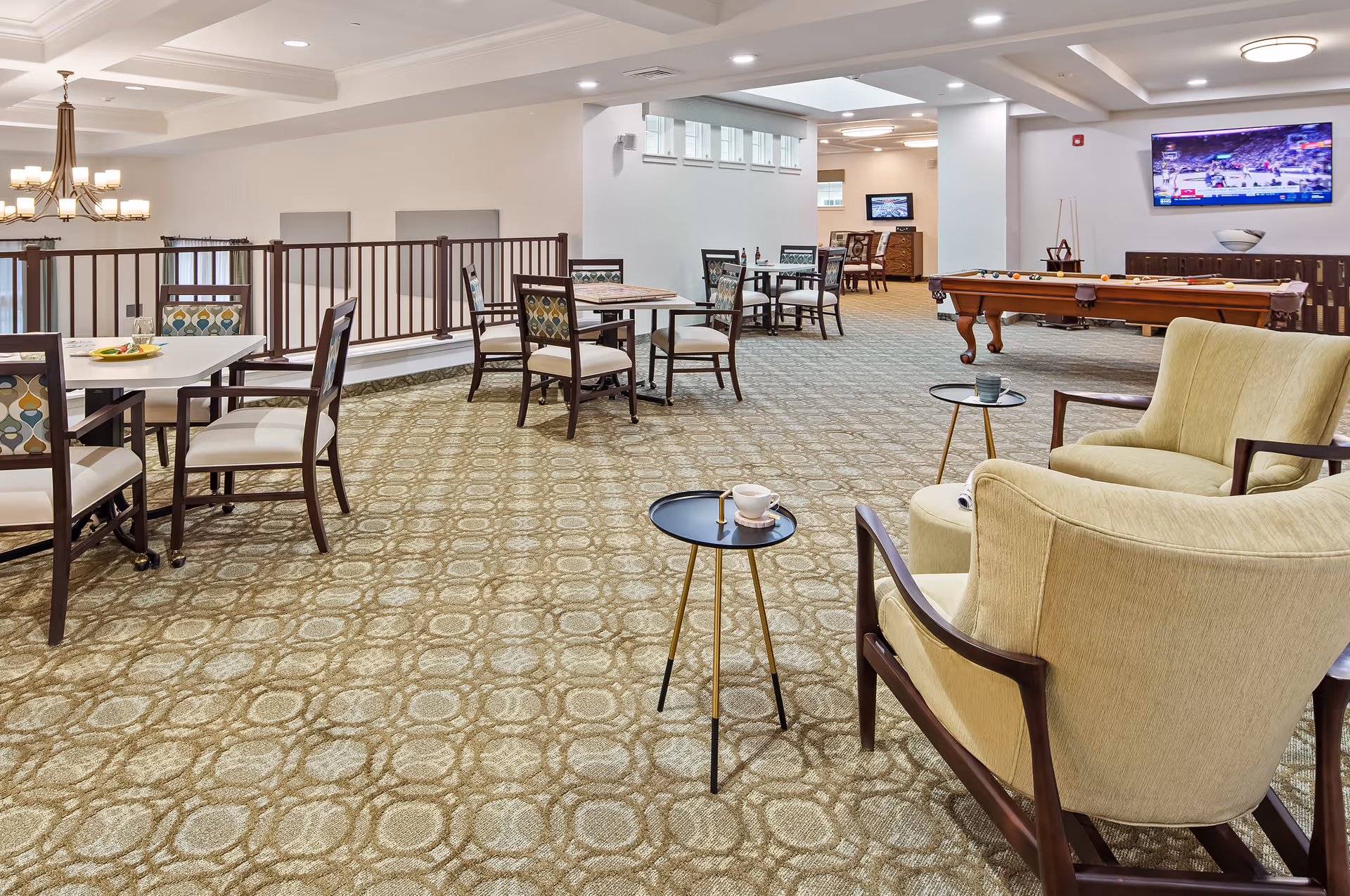 A spacious senior living common area with patterned carpet, several tables and chairs, two comfortable armchairs with small round side tables, a pool table, and two wall-mounted televisions showing sports games.