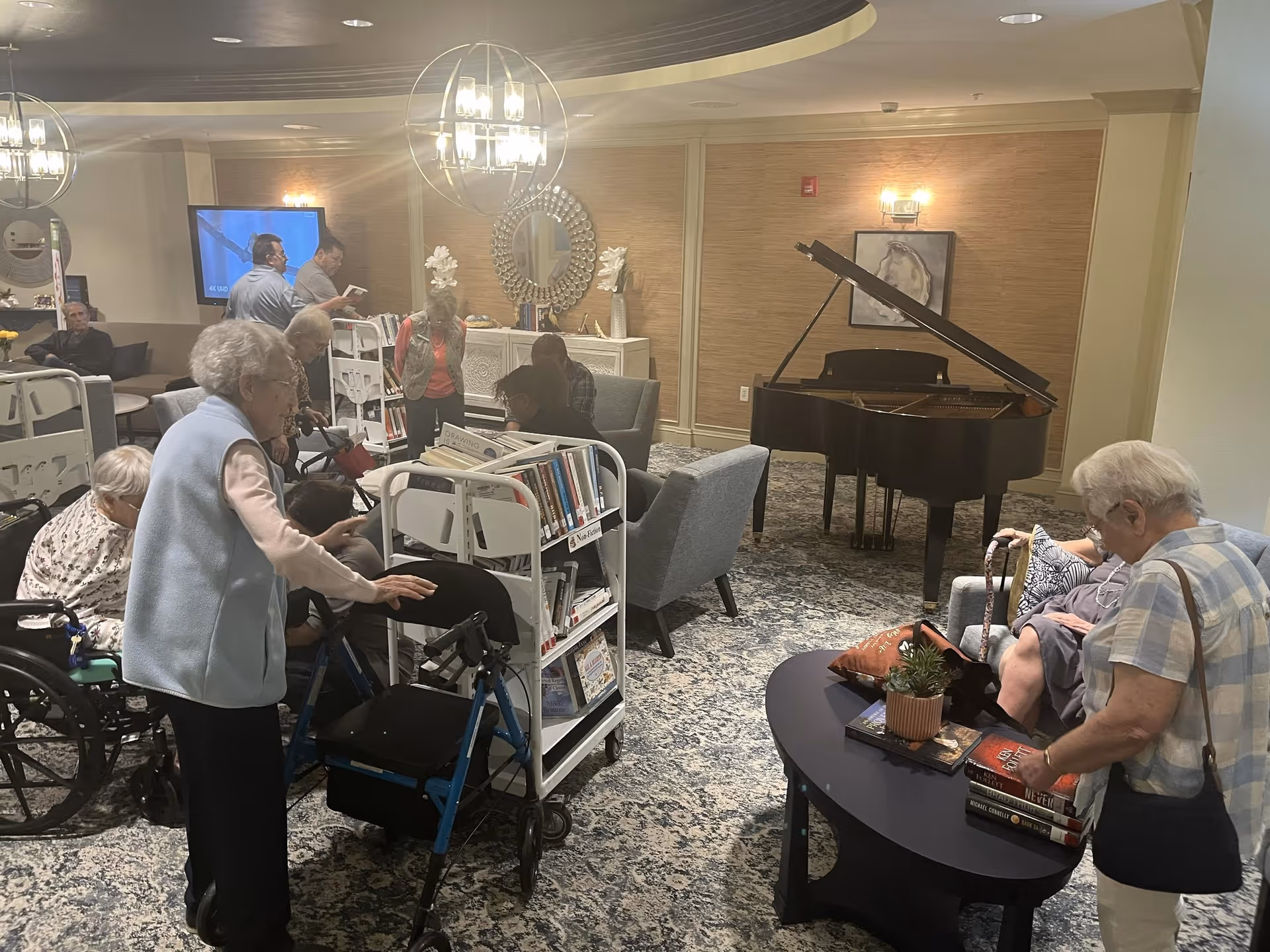 A group of elderly people in a common area of a senior living facility. Some are seated, others are standing or using walkers. There are bookshelves with books, a grand piano, comfortable chairs, and a round table with books and a small plant. The room has warm lighting and decorative elements including a mirror and wall art.