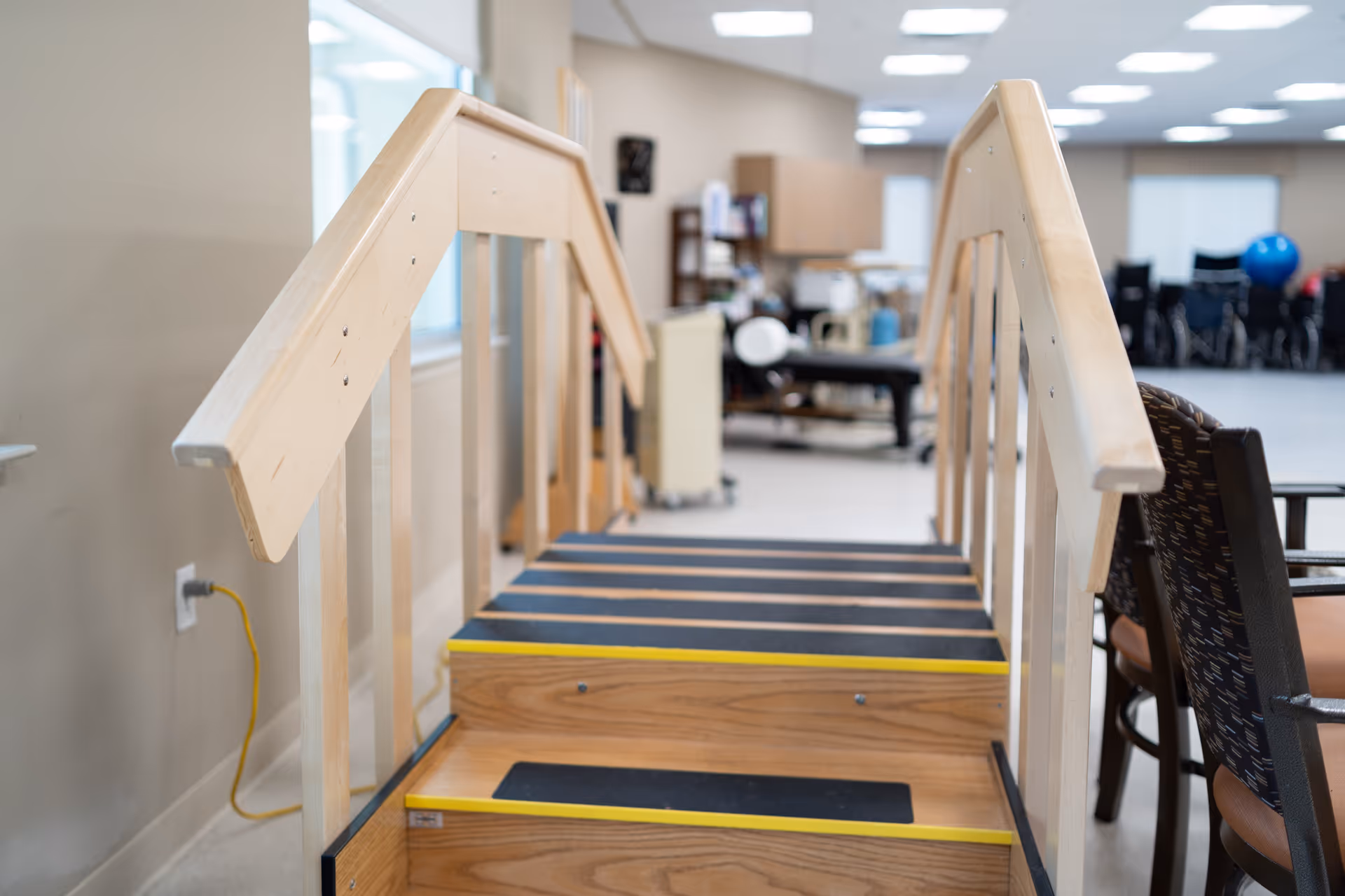 Indoor physical therapy area with a wooden staircase featuring black anti-slip strips and yellow edges, handrails on both sides, and exercise equipment and chairs visible in the background.