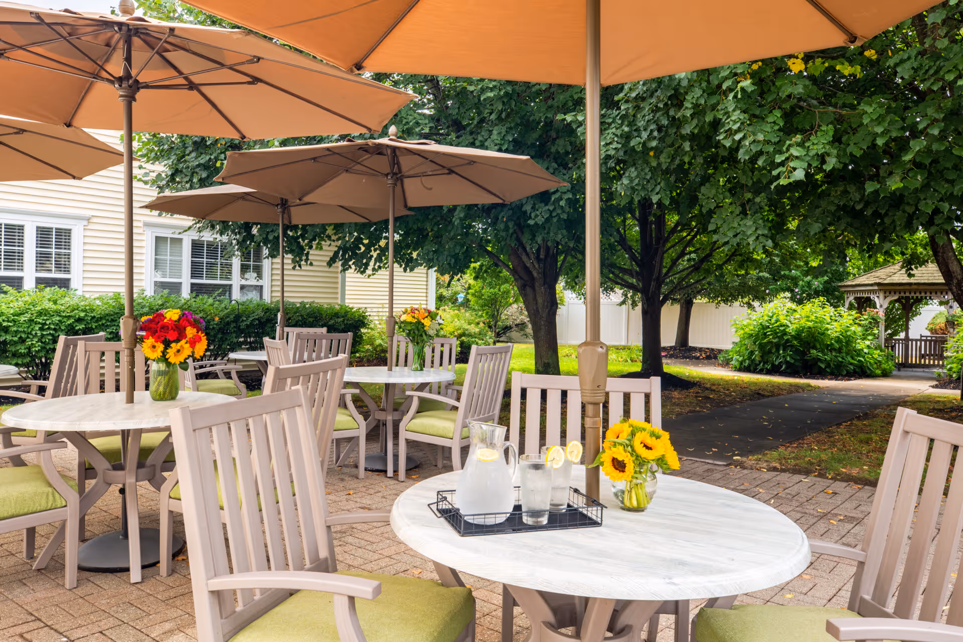 Outdoor patio with round tables and umbrellas, cushioned wooden chairs, flower centerpieces, and a gazebo among trees.