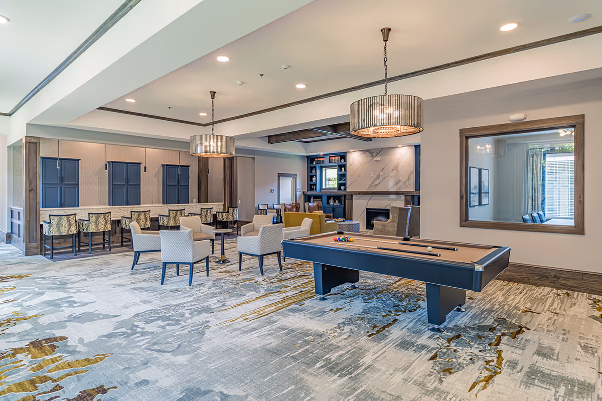 A spacious senior living common area featuring a pool table in the foreground, several upholstered chairs arranged around small tables, a bar counter with high chairs, and a cozy seating area with armchairs near a fireplace and built-in shelves. The room is well-lit with modern pendant lights and recessed ceiling lights, and has a large window with wooden trim on one wall.