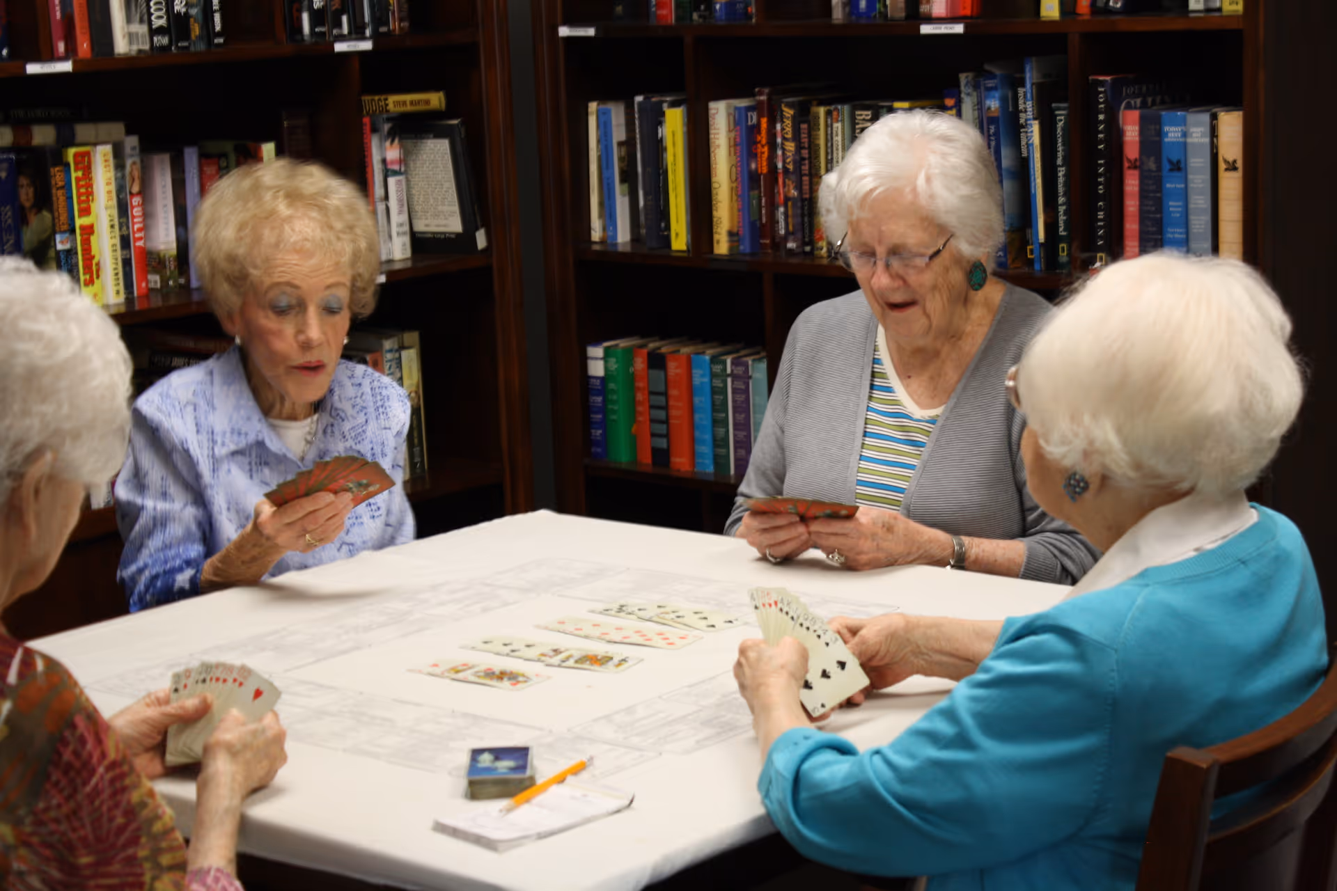 Four elderly women sitting around a table playing cards in a room with bookshelves filled with books in the background.